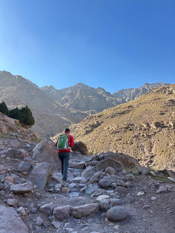 A guide in a red jacket climbing a rocky path towards the Toubkal massif