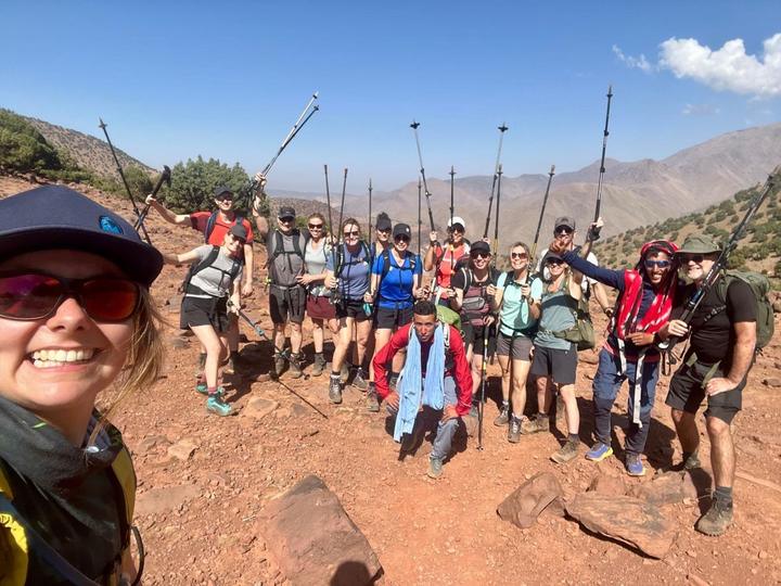 A large group of trekkers and their guide raising hiking poles on a red mountain trail