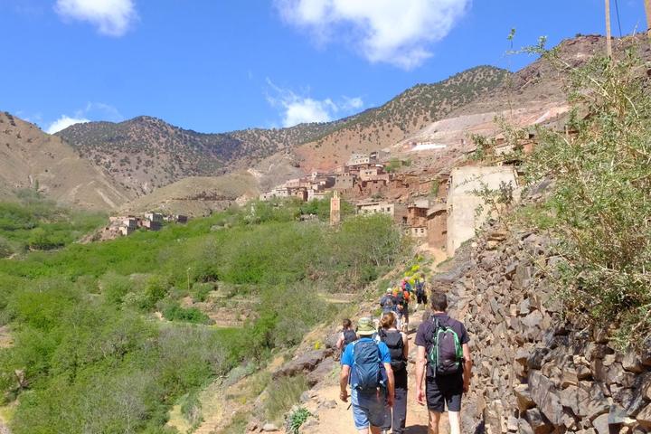 Trekkers walking a village path towards red-earth houses and a mosque tower